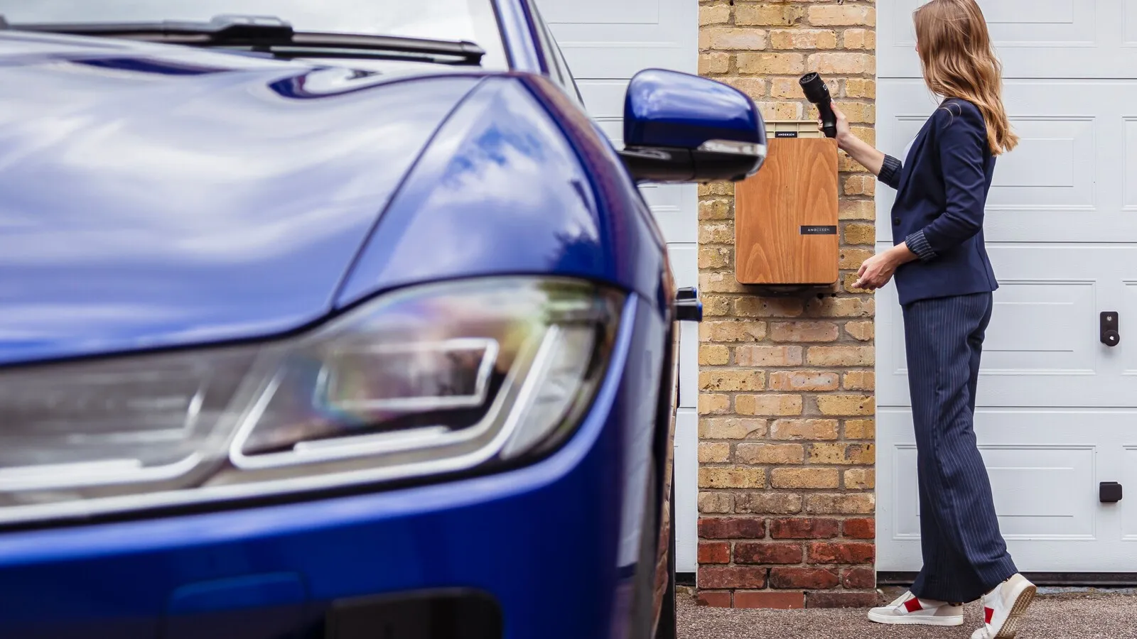 A woman charging an electric vehicle from a wall-mounted charge point on a driveway